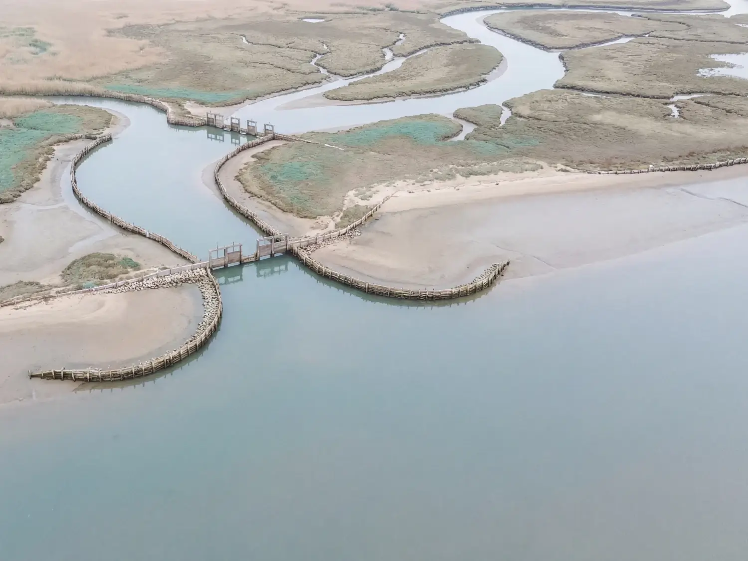 High-angle aerial view of tidal waterways and wetlands with curved wooden barriers controlling water flow, used on the Protirus website to reflect the structured, resilient approach of enterprise cybersecurity experts working in complex environments.